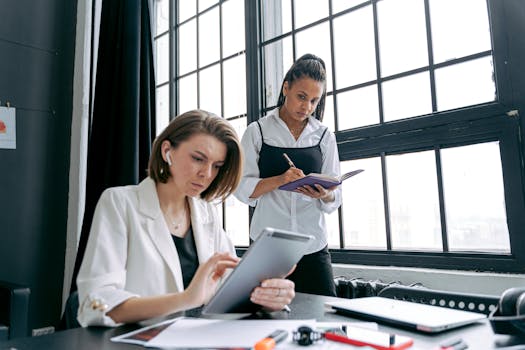 Two professional women working together in a modern office, using technology and documents.