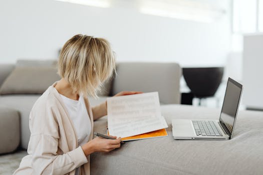 Woman reading documents and working on a laptop at home, creating a cozy and productive environment.