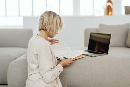 A woman reading while using a laptop in a cozy indoor setting, emphasizing multitasking and relaxation.