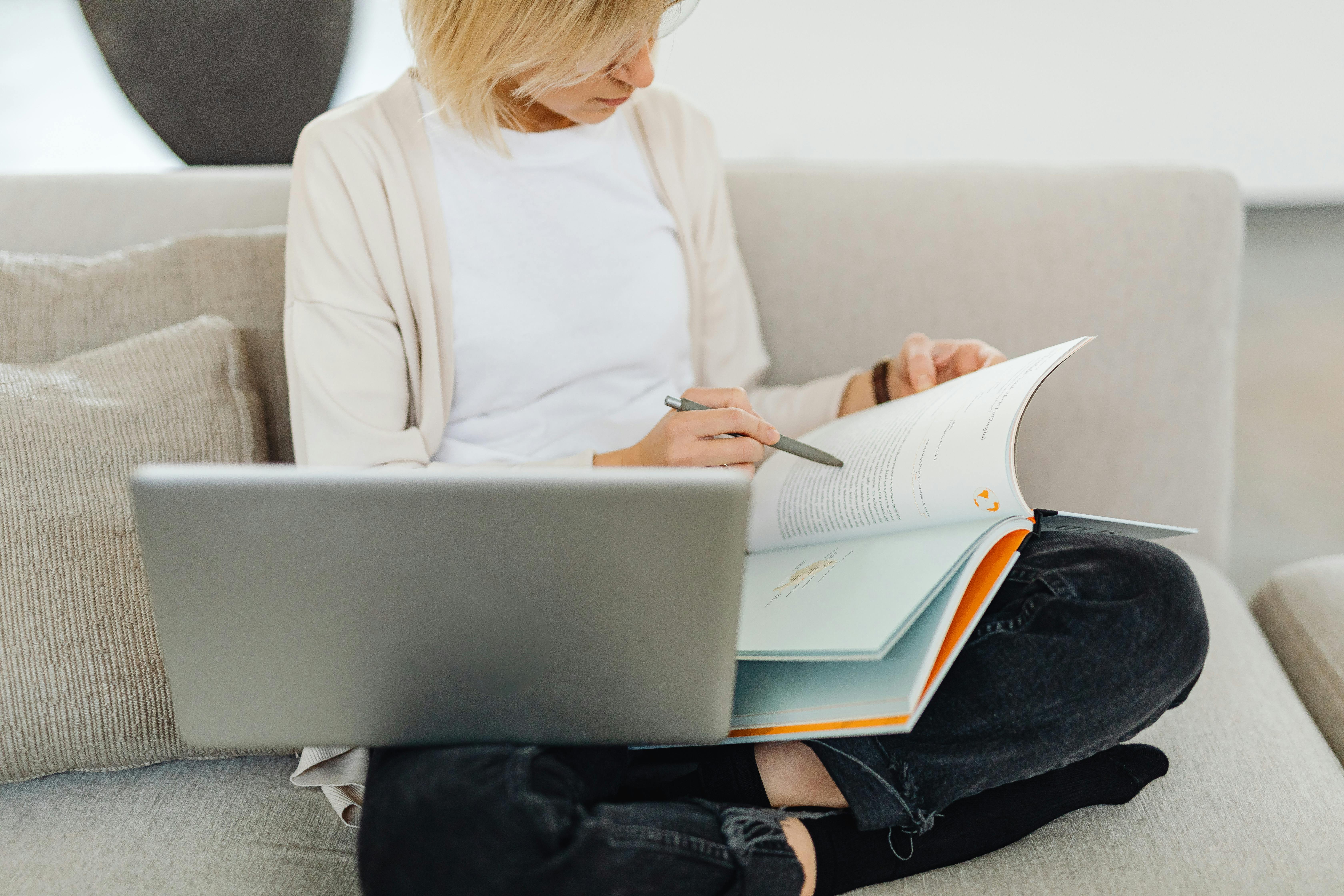 Woman sitting cross-legged on sofa, studying with a textbook and laptop, creating a cozy learning environment.