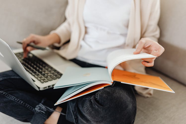 A Person Sitting On A Couch Typing On Her Laptop While Flipping Pages Of A Book