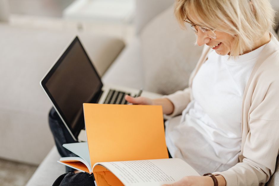 Adult woman with eyeglasses reading a book and using a laptop at home.