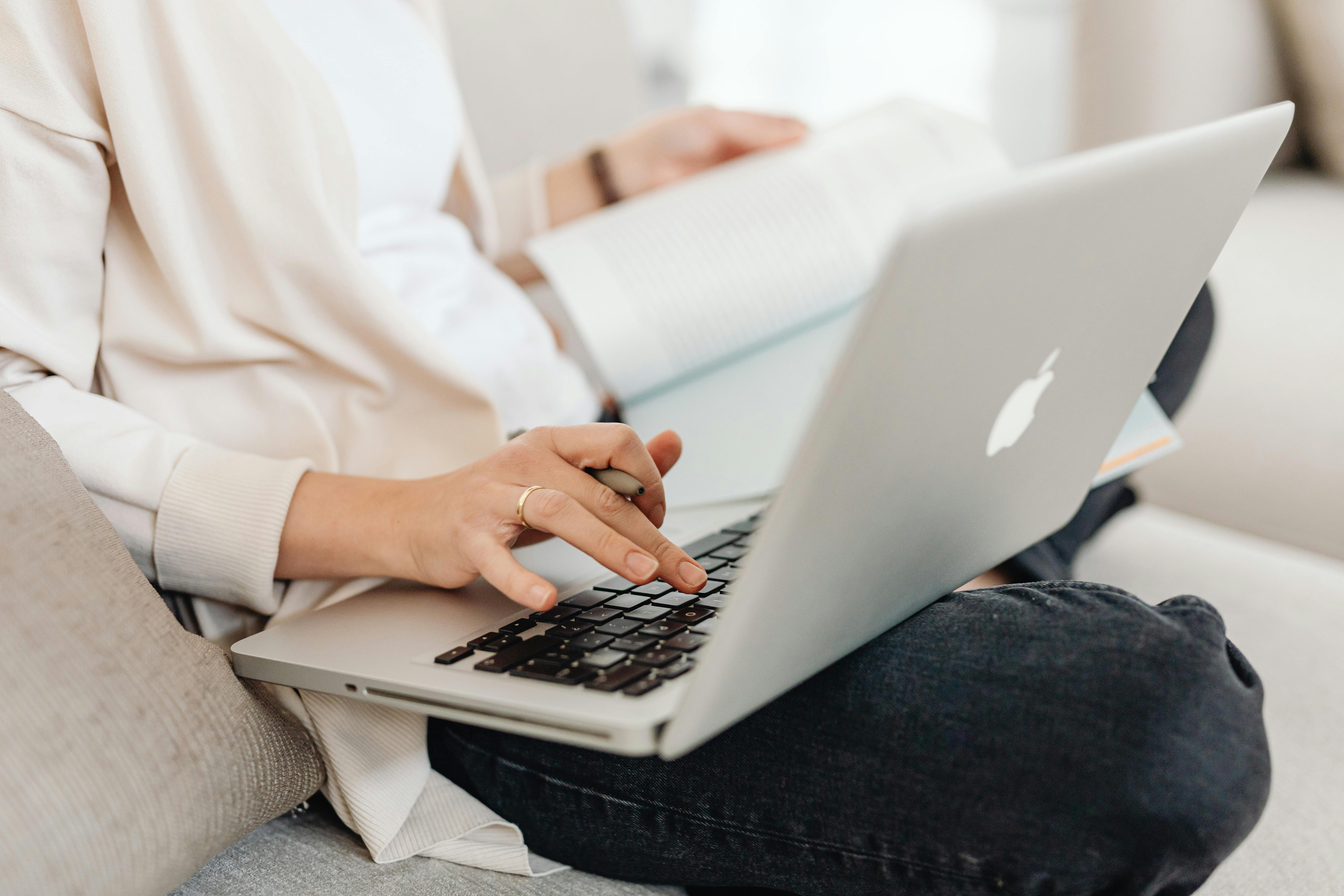 Close-up of a person multitasking, typing on a laptop while holding a book on their lap.