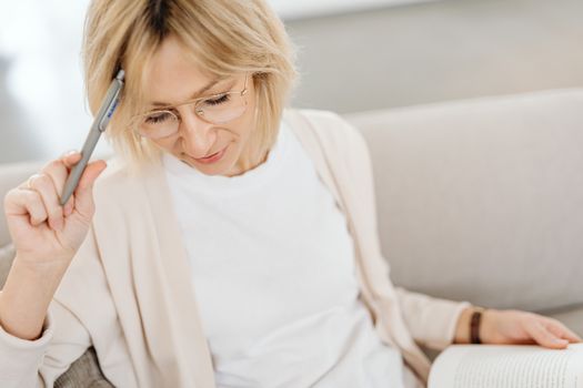 Blonde woman with eyeglasses in deep thought holding a pen while reading a book.