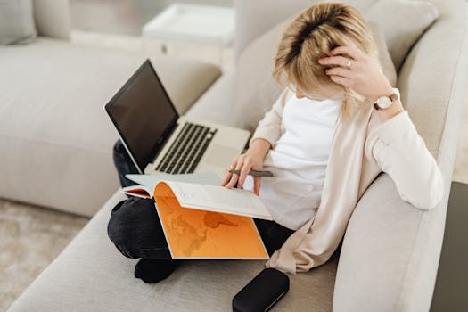 A woman relaxing on a sofa while reading a book and using her laptop, creating a cozy work from home atmosphere.