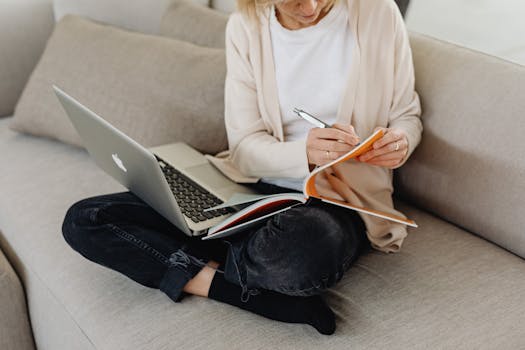 A woman sitting cross-legged on a cozy couch, focused on writing in her notebook with a laptop nearby.