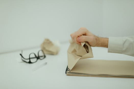 A close-up of a hand crumpling paper on a desk with glasses in the background, evoking frustration.