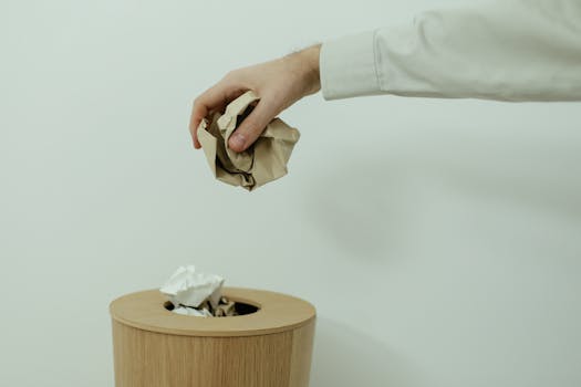 A person's hand disposes of crumpled paper into a wooden trash bin indoors.
