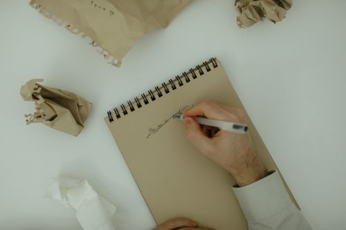 Free Person writing on a notepad surrounded by crumpled paper, symbolizing brainstorming and creativity. Stock Photo