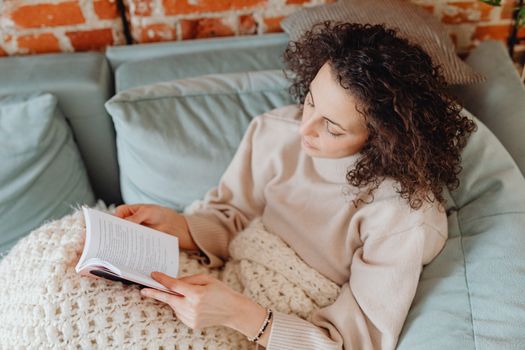 A woman reading a book on a comfortable sofa with a knitted blanket on a relaxed day.