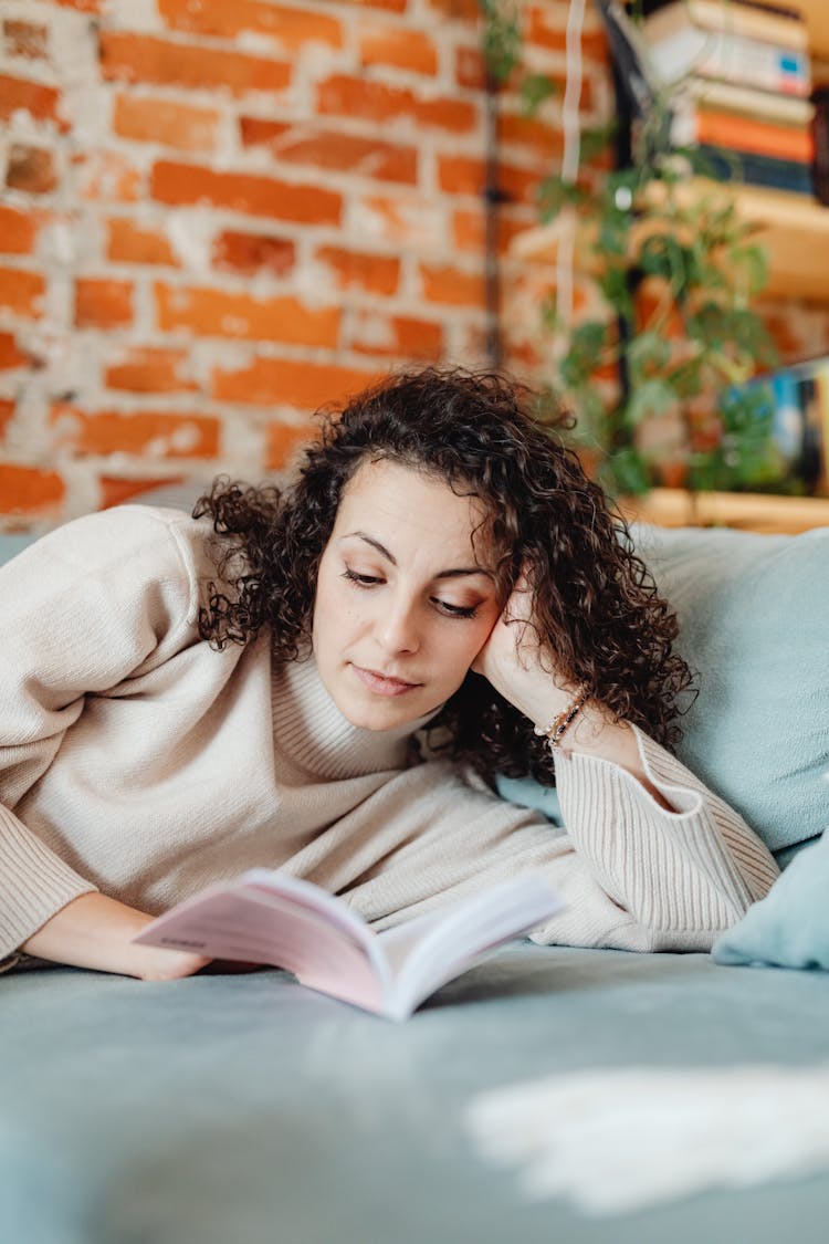 A Woman In Curly Hair Wearing Turtle Neck Sweater Resting On A Couch While Reading A Book