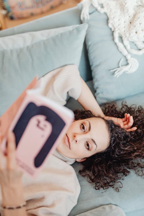 A Woman Lying on a Couch Beside Throw Pillows while Reading a Book