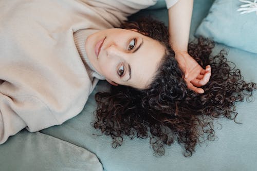 Free A Woman with Curly Hair Wearing Turtle Neck Sweater Lying on a Bed while Looking Afar Stock Photo