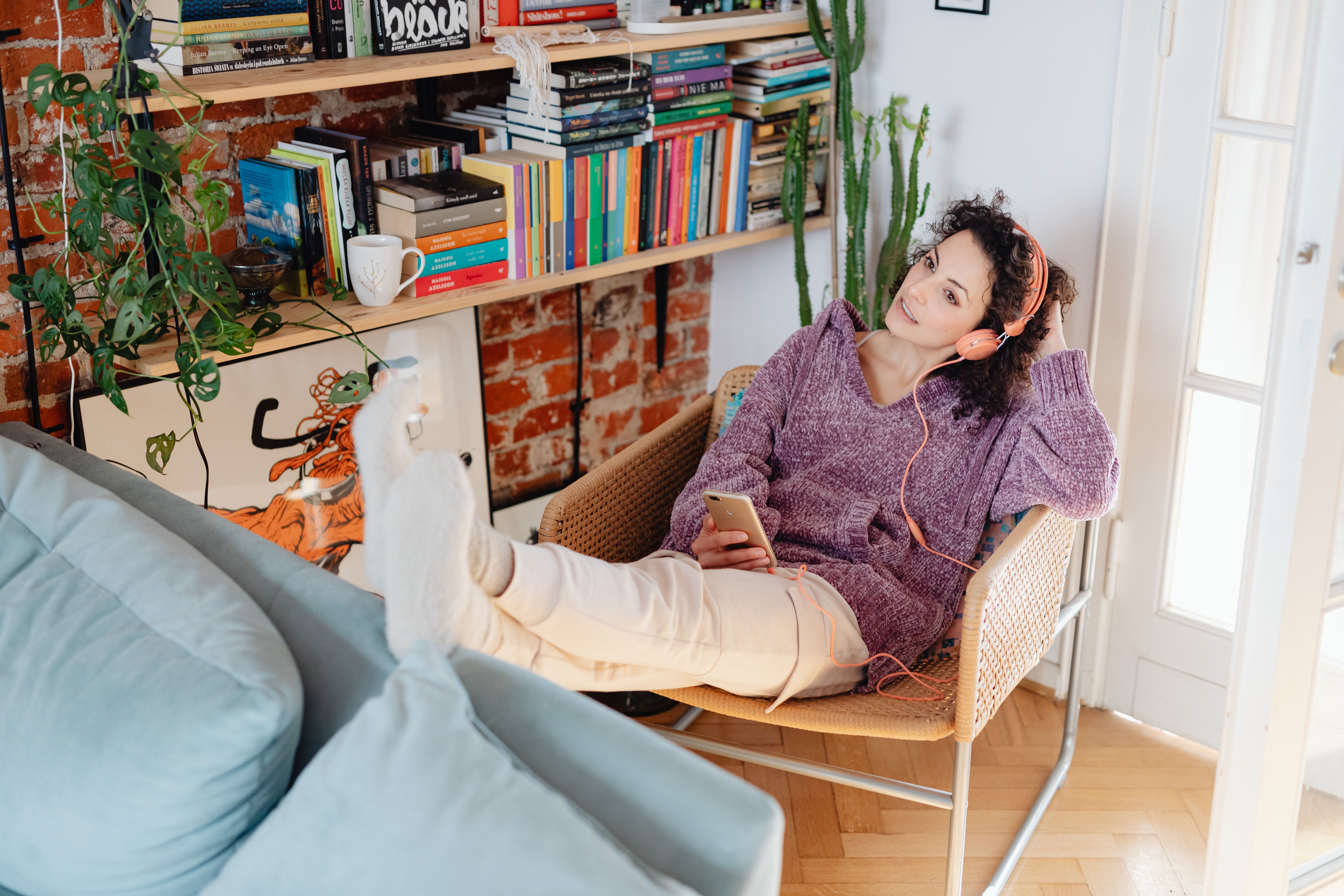 Mulher usando fones de ouvido, relaxando com o smartphone, cercada por livros e aproveitando o tempo livre.