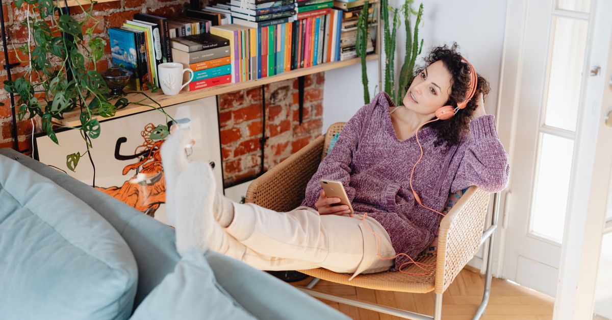 Mulher usando fones de ouvido, relaxando com o smartphone, cercada por livros e aproveitando o tempo livre.