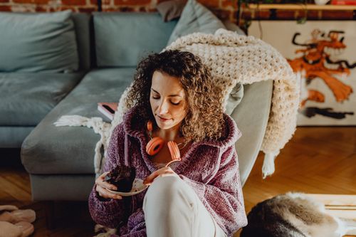Free A woman in a cozy knitted sweater sitting indoors and enjoying a cupcake with headphones around her neck. Stock Photo