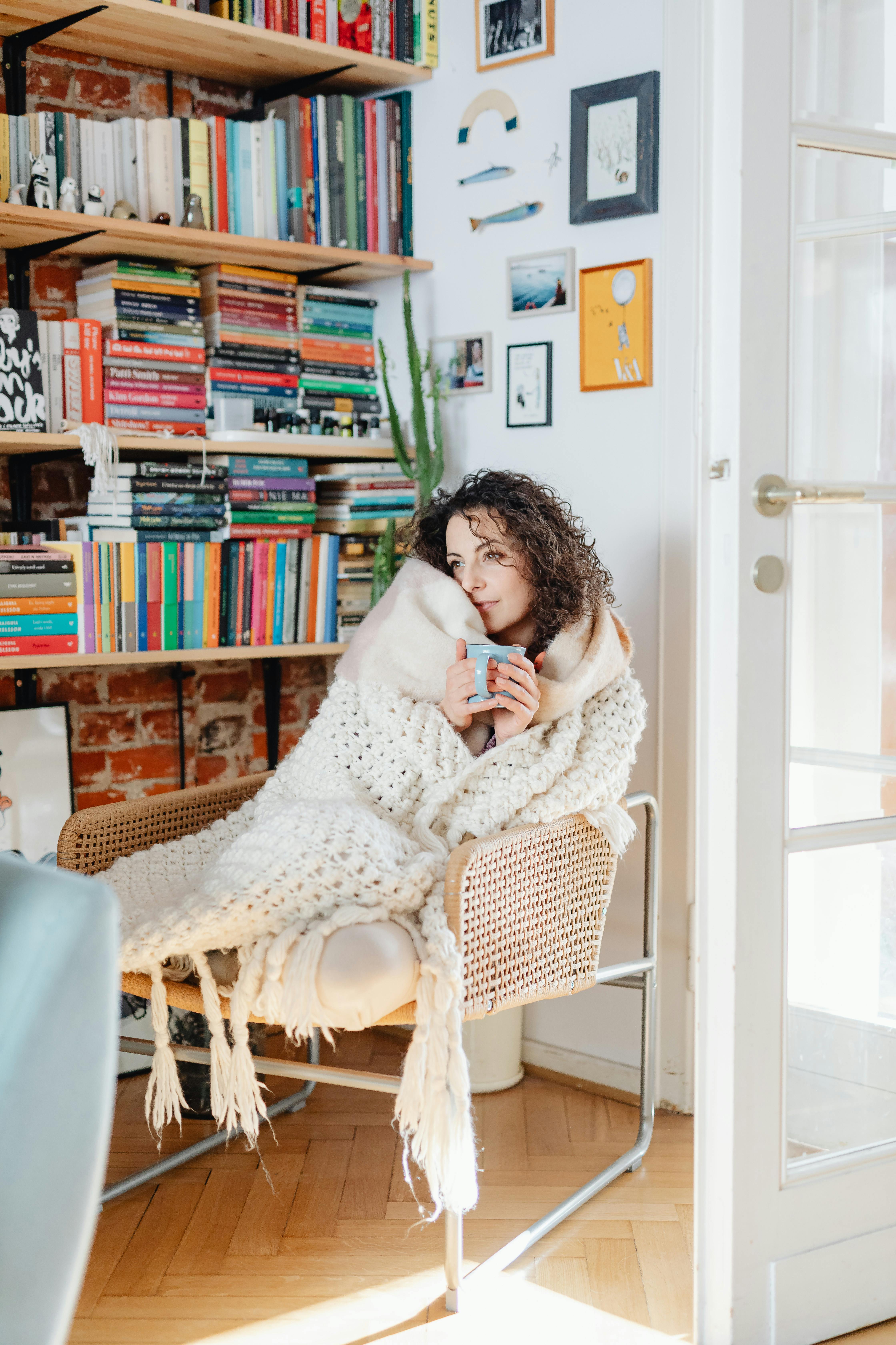 Free A woman wrapped in a blanket enjoying a warm drink in a cozy room filled with bookshelves. Stock Photo