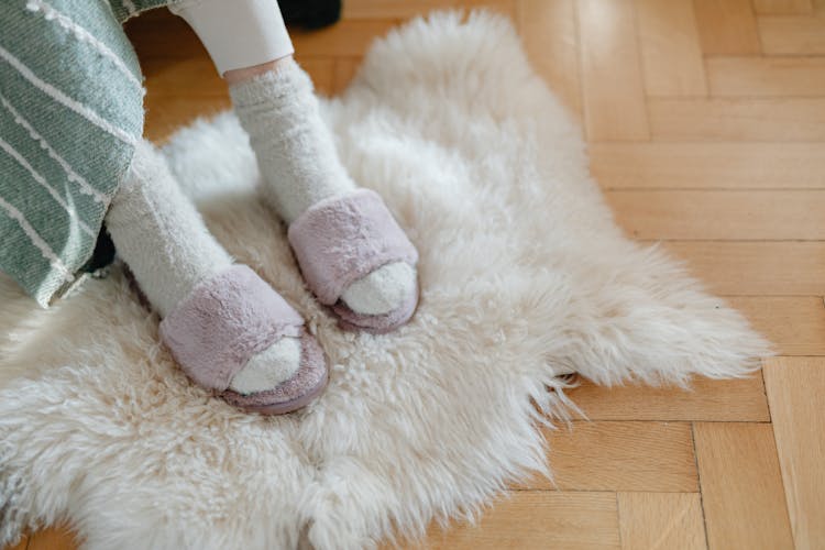 Feet In Warm Terry Socks And Slippers On White Fur Rug 