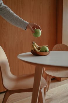 A hand reaches for a green apple in a minimalist kitchen setting with bananas in a wooden bowl.