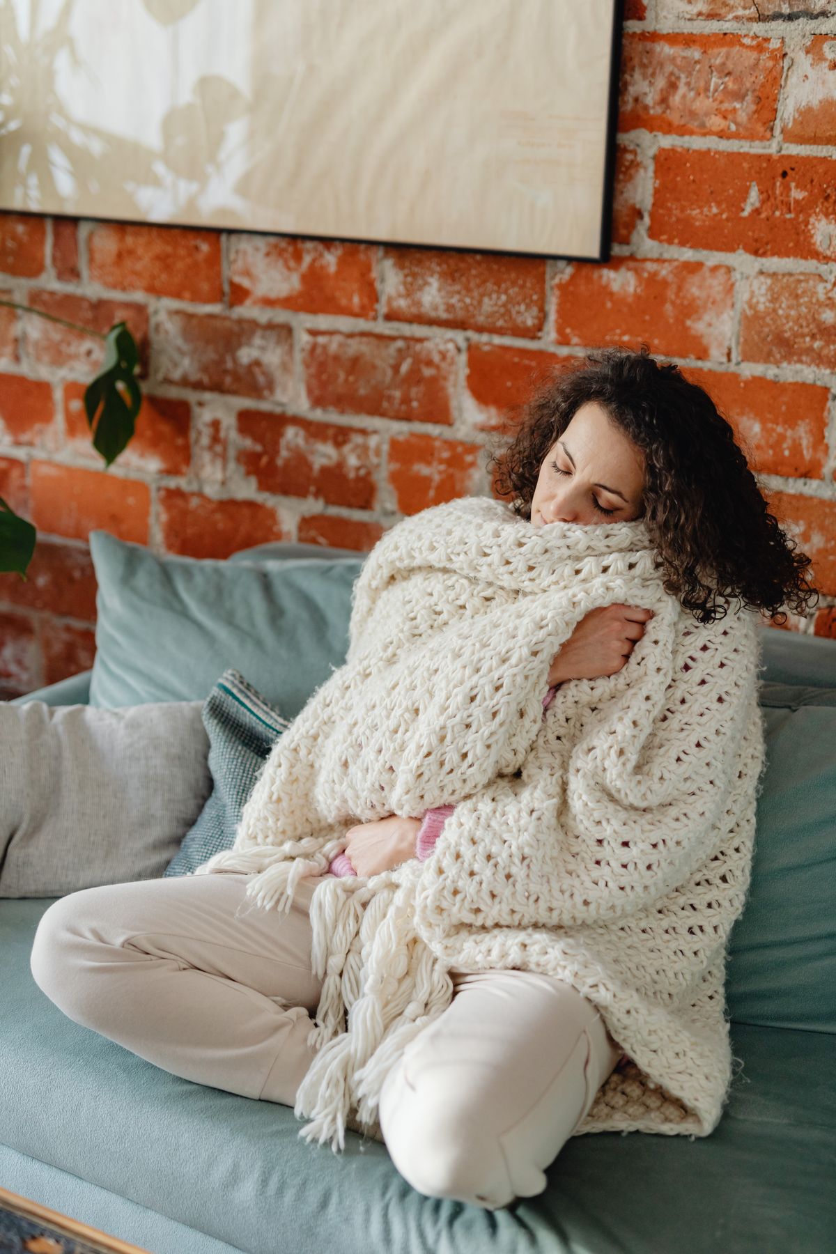 A woman wrapped in a thick knitted blanket on a sofa, holding her hands against her middle