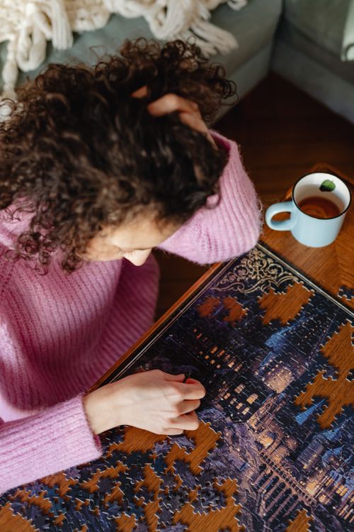 Free A woman in a pink sweater solves a complex puzzle at a wooden table near a cozy couch. Stock Photo