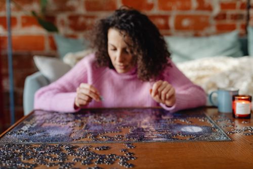 Free A woman in a pink sweater assembling a jigsaw puzzle on a table in a cozy setting. Stock Photo