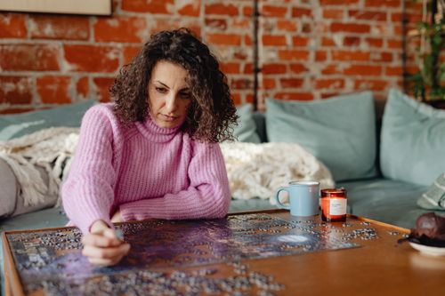Free Woman in pink sweater solving a jigsaw puzzle on wooden table indoors. Cozy atmosphere. Stock Photo