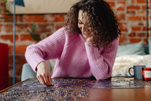 Free Woman with curly hair and pink sweater solving a jigsaw puzzle indoors, exuding a relaxed and cozy vibe. Stock Photo