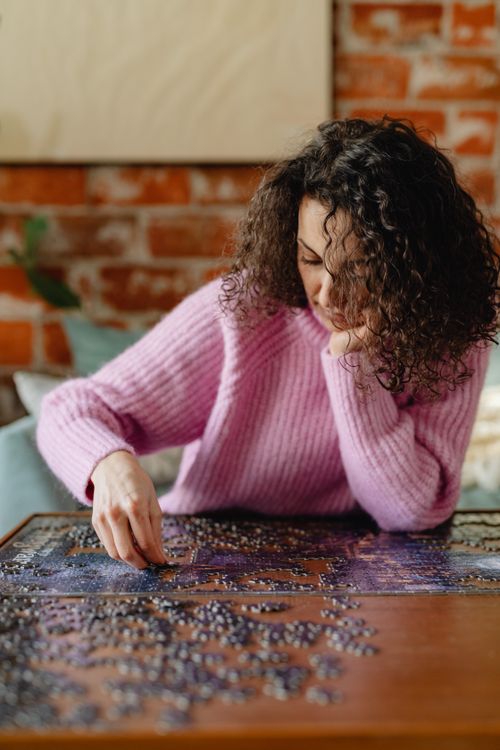 Free A woman in a pink sweater thoughtfully works on a jigsaw puzzle indoors, creating a cozy atmosphere. Stock Photo