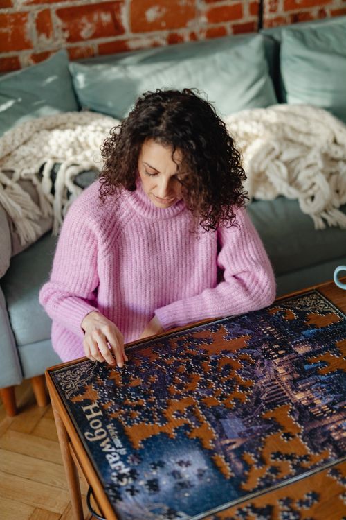 Free Woman in pink sweater assembling a jigsaw puzzle on a wooden table, capturing a moment of relaxation and joy. Stock Photo