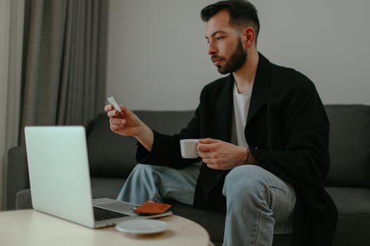 A man enjoying coffee while checking his phone and using a laptop in a cozy living room setting.
