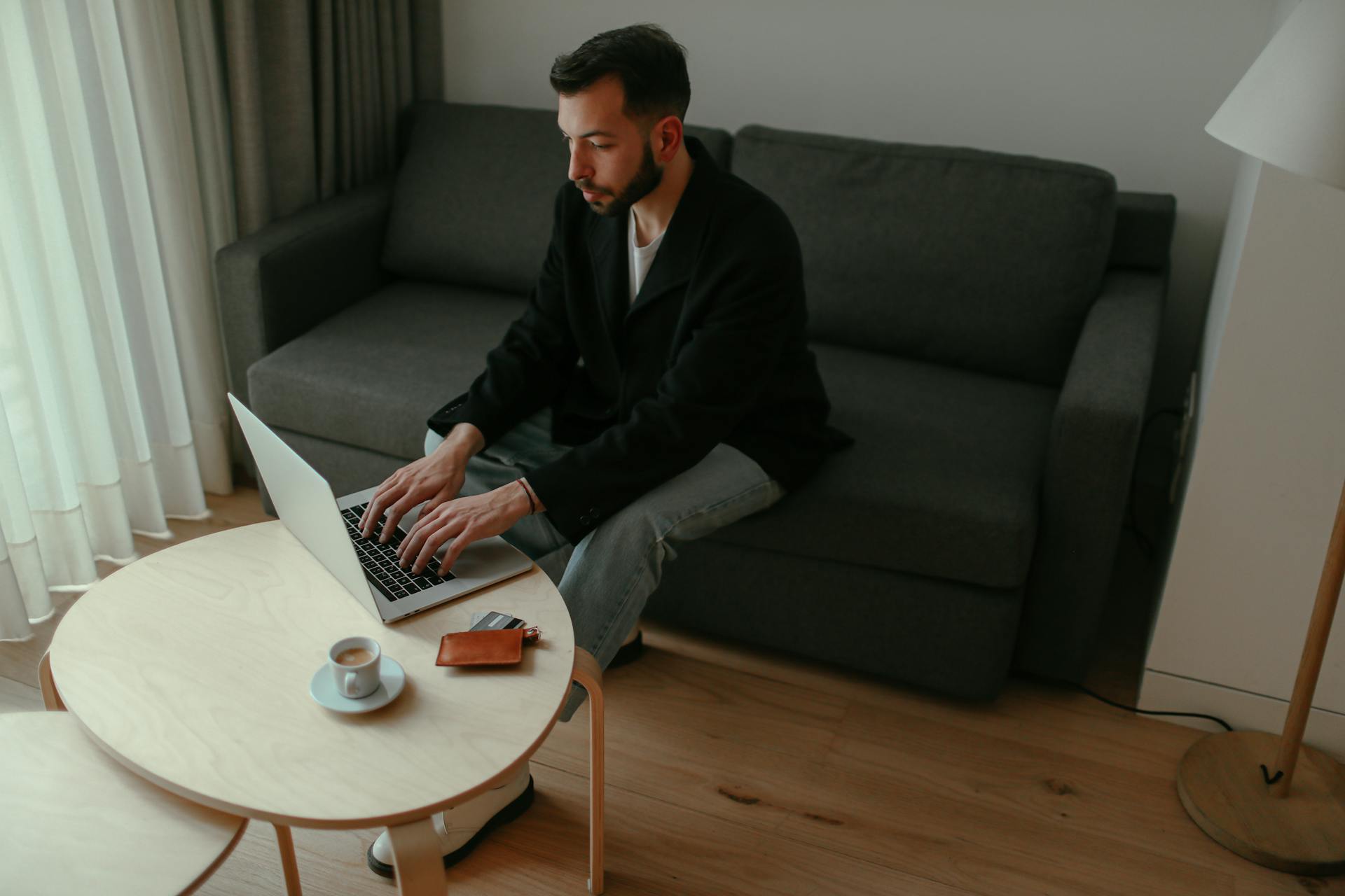 Man sitting on couch typing on laptop, online shopping at home with coffee and wallet nearby.