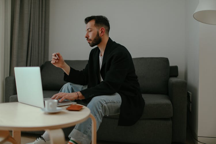 Man In Black Blazer And Denim Pants Sitting On A Couch While Using A Laptop