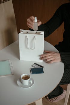 Person holding a dropper above a shopping bag with credit cards and tablet on table.