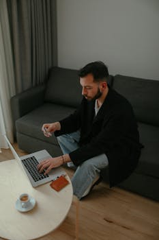 Adult man using laptop and holding credit card, sitting on a sofa for online shopping.
