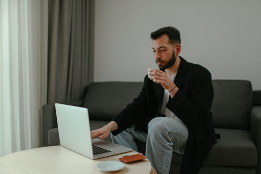Adult man sitting on couch working on laptop with coffee cup in hand.