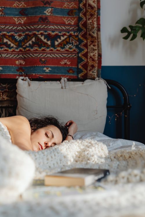 Free A woman peacefully asleep on a cozy bed with a patterned tapestry and soft lighting. Stock Photo