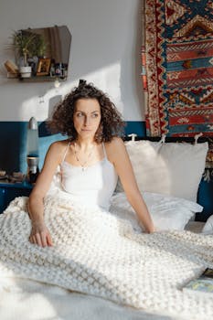 Woman with curly hair enjoying morning sunlight in a cozy bedroom setting.
