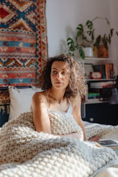Woman sitting on bed wrapped in a knitted blanket, enjoying a cozy morning sun.