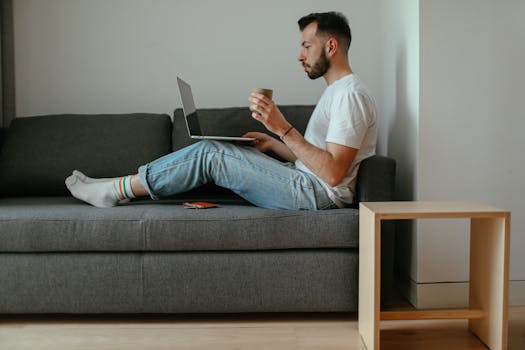 Man sitting on a couch with a laptop and coffee, enjoying a cozy indoor setting.