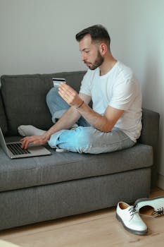 A man sitting on a sofa using a laptop for online shopping with a credit card in hand.