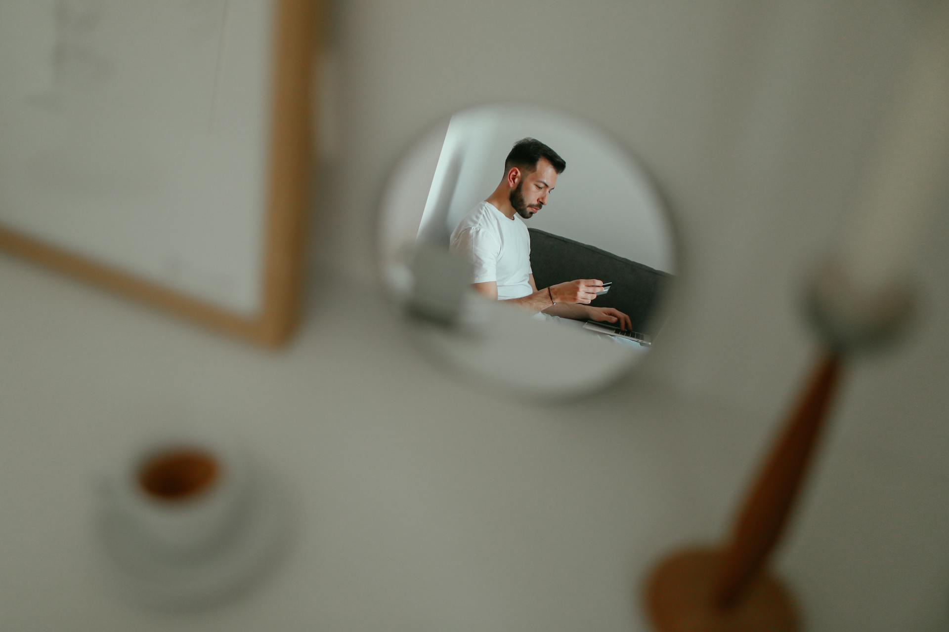 A man reflected in a mirror holding a credit card, engaged in online shopping from his living room.