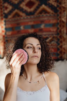 A woman with curly hair uses a pink facial sponge in a relaxing setting.