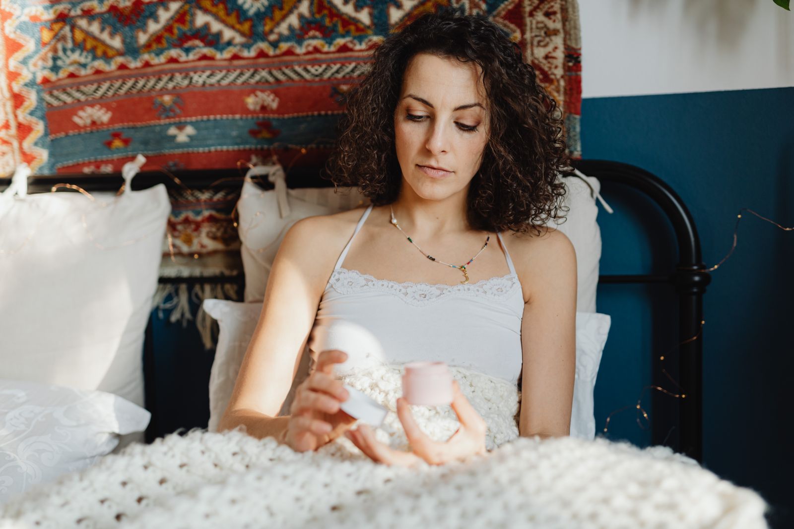Curly-haired woman sitting in bed in warm sunlight, holding a small pink jar of cream