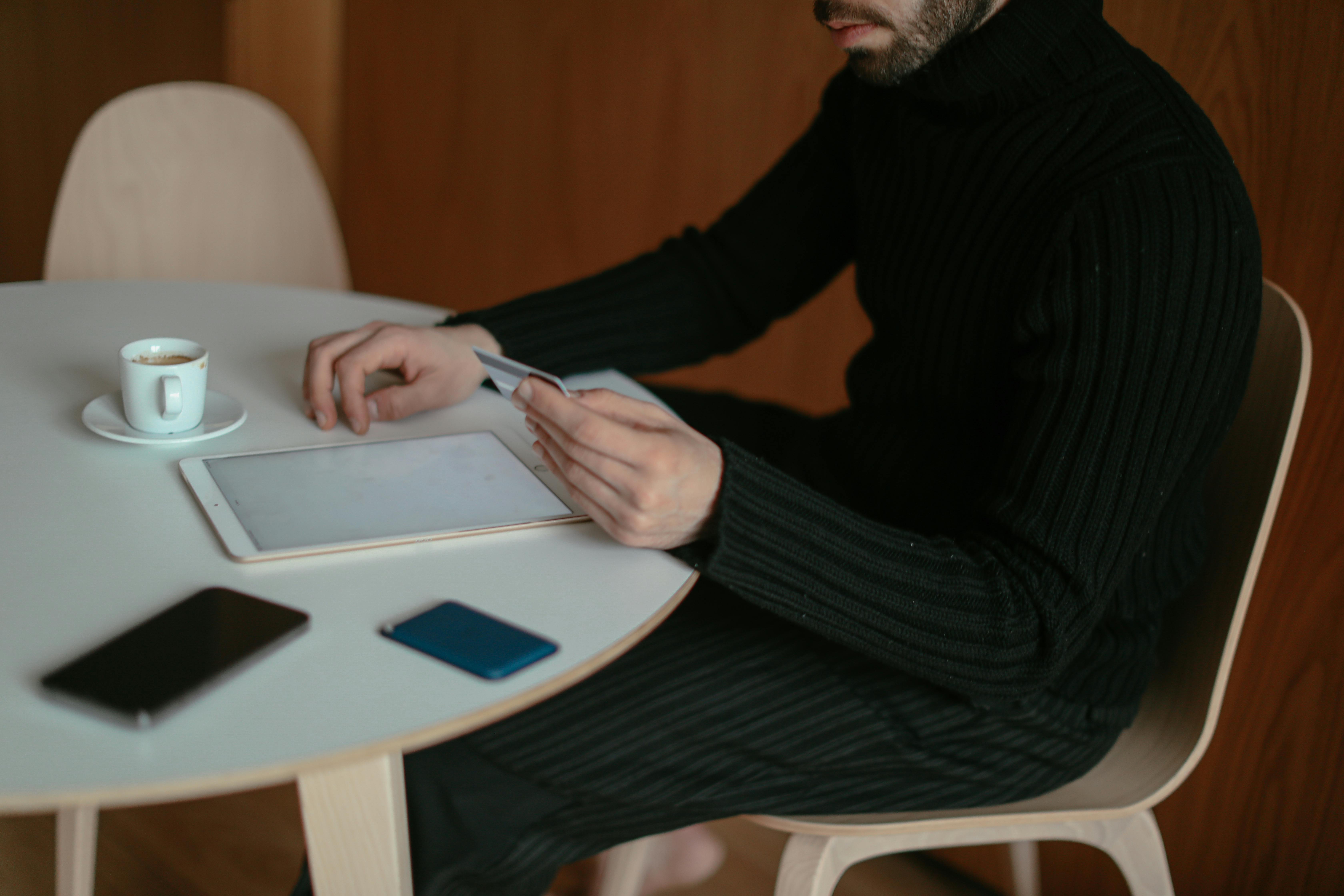 Adult man using tablet and credit card to shop online at home, sitting by table with coffee.