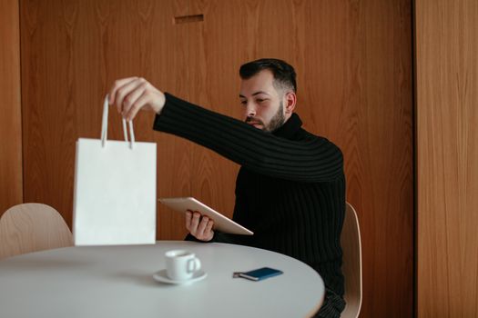 A bearded man in a cafe with a digital tablet holding a white shopping bag.