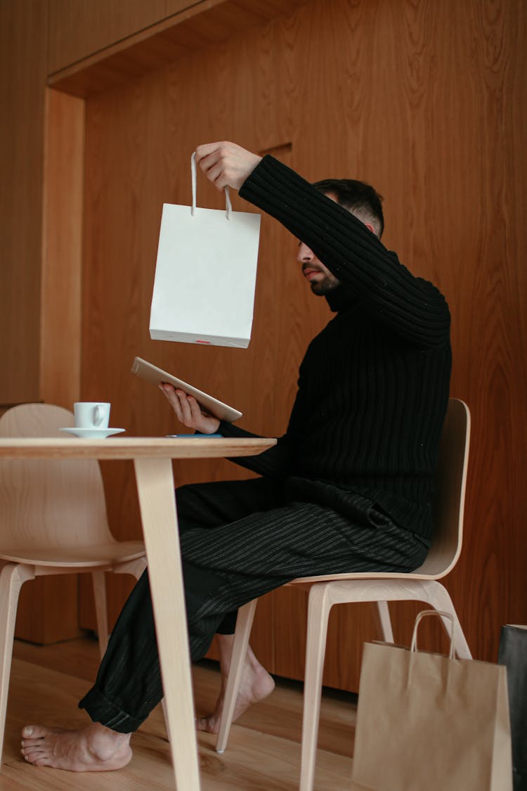 Man In A Wooden Interior Sitting At A Table And Holding A White Paper Bag And Tablet