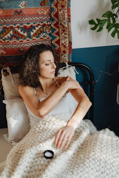 Curly-haired woman applies skincare cream while lying in a cozy, bohemian-style bedroom.