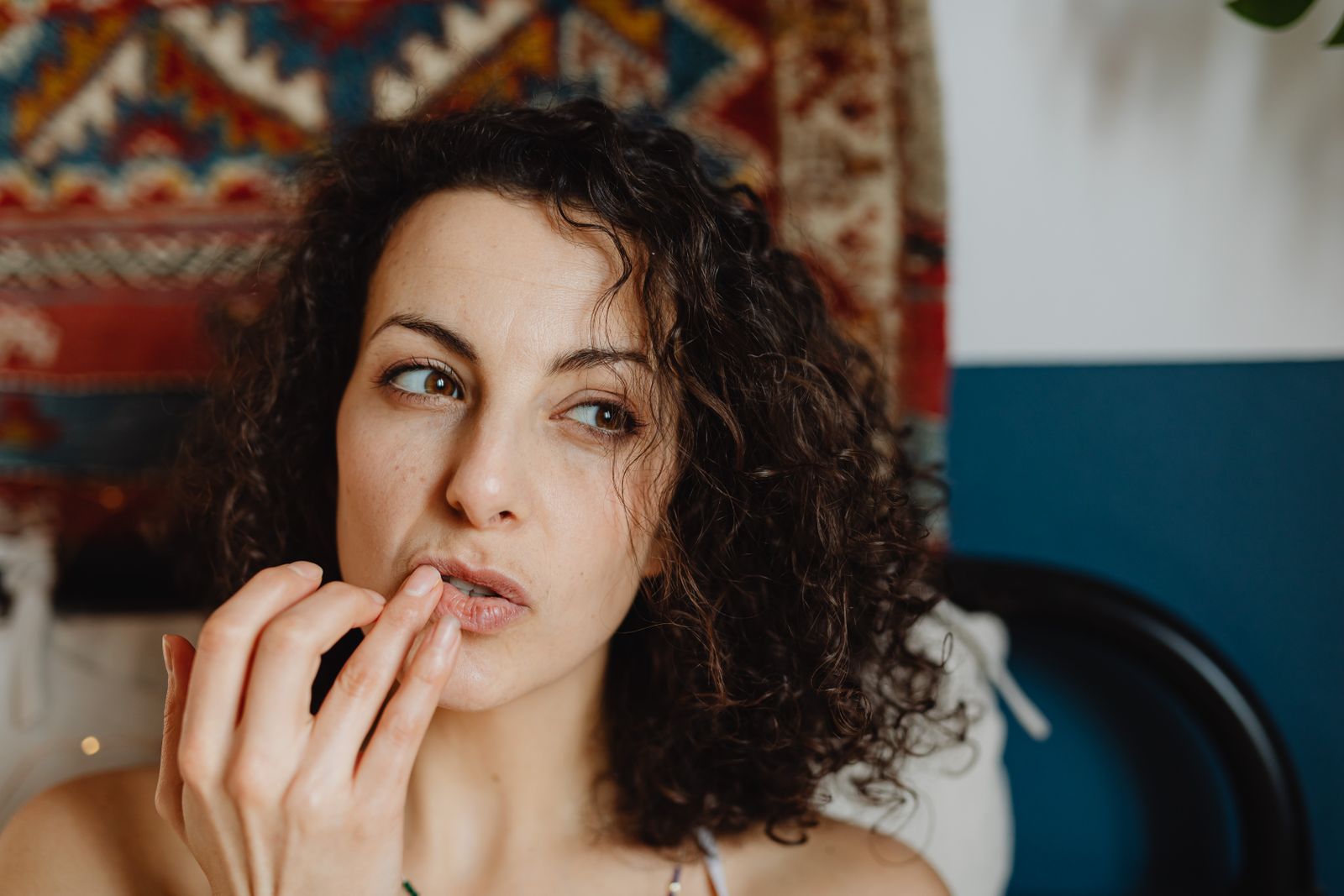 Woman with curly dark hair presses her fingertips to her lips in a cozy room with a patterned wall rug