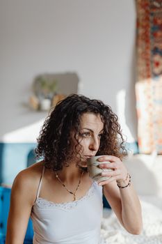 Woman with curly hair drinking coffee in a cozy indoor setting.